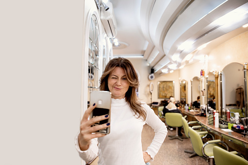 Woman making Selfie Picture using Mobile Phone at Hairdresser Salon. Beautiful Brunette Female with Long Hair sending video message. Copy space