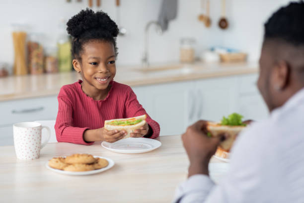 istock Adorable black girl looking at her dad while eating sandwich 1384855573