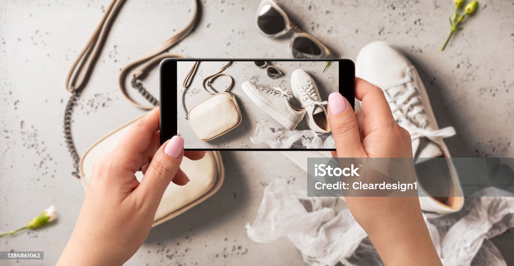 Woman taking photo of spring fashion accessories with smartphone. Influencer and social media. Woman taking photo of white leather sneakers shoes, handbag and sunglasses with smartphone. Blogger, influencer or stylist capturing spring fashion accessories for social media. Grey background. Second Hand Sale Stock Photo