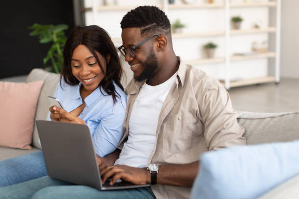 istock Happy black couple sitting on couch, using cellphone and pc 1384861432