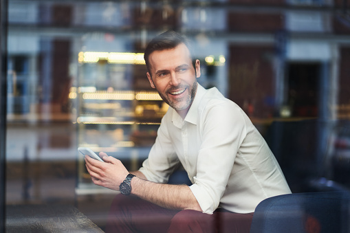 Smiling mid adult businessman sitting in cafe holding smartphone looking through the window