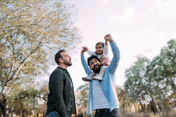 Gay couple with their young daughter enjoying a day in a park. The father raises the arms of the daughter smiling at her. Gay couple with their young daughter enjoying a day in a park. The father raises the arms of the daughter smiling at her. homosexual couple stock pictures, royalty-free photos & images