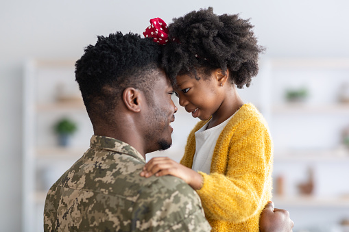 Portrait Of Black Cute Little Girl Embracing With Her Military Father, African American Male Soldier In Camouflage Uniform Cuddling His Daughter, Bonding With Female Child At Home, Side View, Closeup