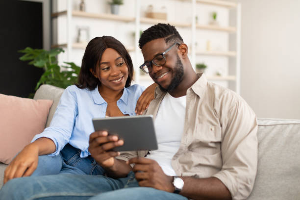 istock Lovely African American couple sitting on sofa, using tablet 1384866560