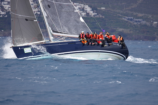 Bodrum,Mugla, Turkey.27 February 2022: sailor team driving sail boat in motion, sailboat wheeling with water splashes, mountains and seascape on background. Sailboats sail in windy weather in the blue waters of the Aegean Sea, on the shores of the famous holiday destination Bodrum. Men and women on a sailboat in a very cold weather continue to cruise. Sailing crew on sailboat during regatta. Sailing in the wind through the waves at the Sea. Close up of sailing boat, sail boat or yacht at sea with white sails.