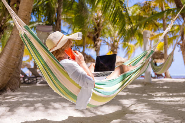Woman lying in hammock on beach and working on laptop and speaking on phone stock photo