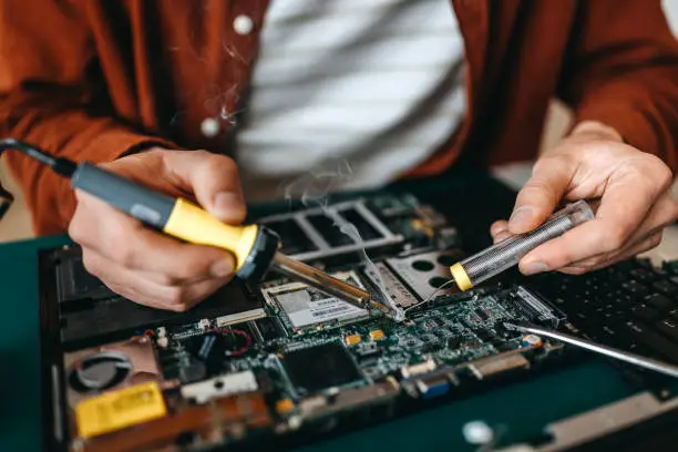 Midsection of young man repairing damaged mother board with soldering iron and tool on table at home