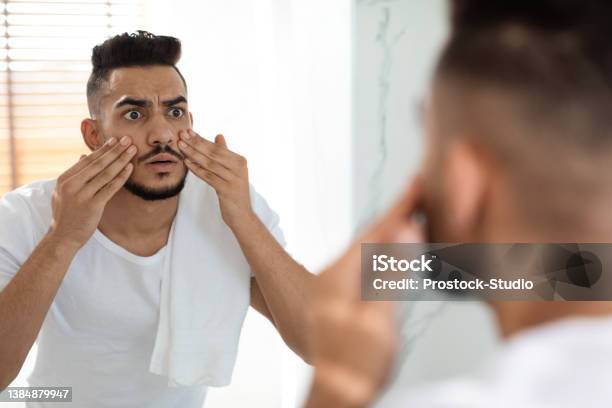 Dull Skin Concept Worried Arab Guy Looking At Mirror And Touching Face Stock Photo - Download Image Now