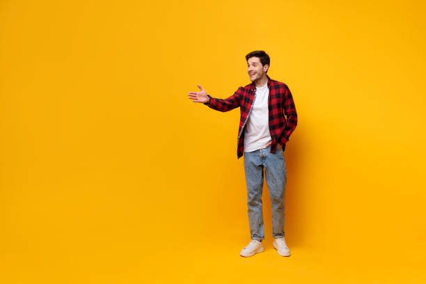 Portrait of happy young man posing at yellow studio Nice to Meet You. Handsome young guy outstretching hand to the side offering handshake or showing free copy space and smiling on yellow studio background, greeting someone, full body length banner casual handshake stock pictures, royalty-free photos & images