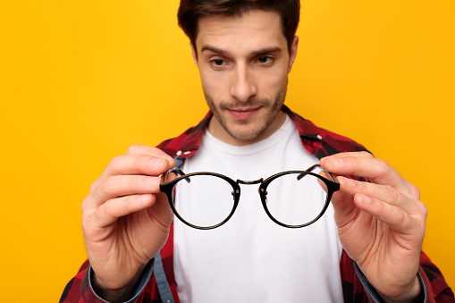 Closeup portrait of young man holding and looking at eyeglasses, showing spectacles close to camera, selective focus. Millennial guy standing isolated on yellow studio blurred background wall
