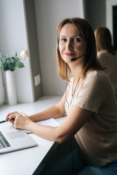 istock Vertical portrait of smiling female operator talking using headset and consulting client sitting at window sill with laptop from home office 1384887870