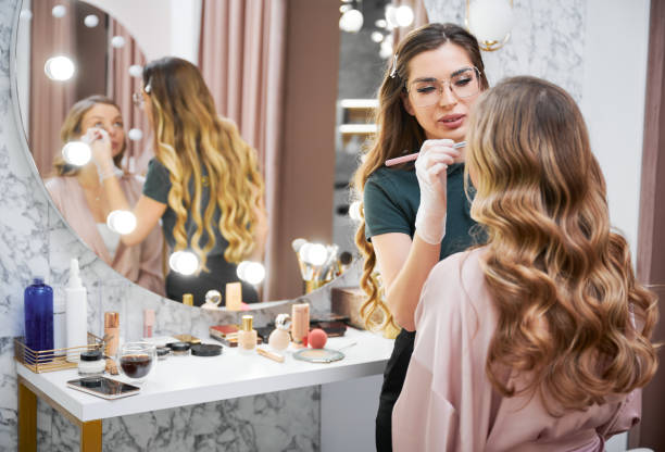 Female makeup artist doing makeup on woman in beauty salon. Female stylist doing professional makeup for client in beauty salon. Young woman sitting at dressing table and looking in the mirror while makeup artist applying concealer with cosmetic brush. makeup artist stock pictures, royalty-free photos & images