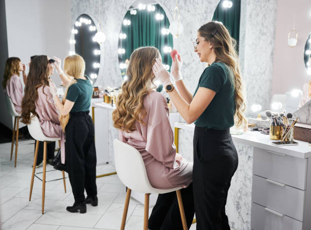 Makeup artists working with clients in beauty salon. Young woman with long curly hair sitting at dressing table while female stylist applying foundation with sponge. Beauty specialists doing professional makeup for clients in modern salon. salon mirror stock pictures, royalty-free photos & images