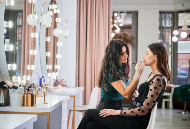 Smiling makeup artist doing makeup for woman in beauty salon. Makeup artist doing professional makeup in visage studio. Stylish woman sitting at dressing table while female beauty specialist in sterile gloves applying lipstick on client lips with cosmetic brush. backstage stock pictures, royalty-free photos & images