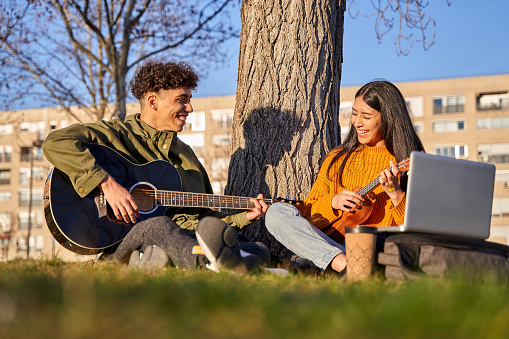 Young latin couple sitting at tree playing ukulele and guitar singing a song. happy feeling/relaxing concept