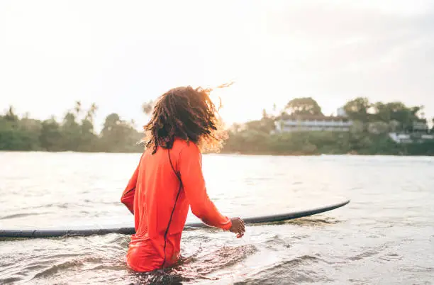 Photo of Black long-haired teen boy with a surfboard ready for surfing. He walking into Indian ocean waves with magic sunset background. Extreme water sports and exotic countries concept.