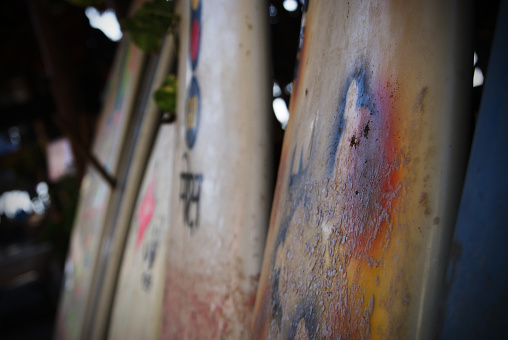 A line of white surfboards stand propped against a wall in a dimly lit shack. Wax and sand can be seen on their surface.