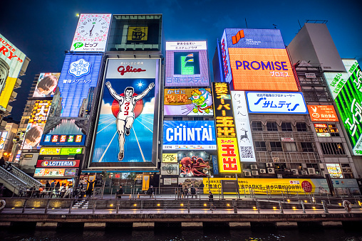 Osaka, Japan - March 28, 2019:  Tourists and locals walk below the famed advertisements lining Dotonbori Canal at night. The district is one of Osaka's primary tourist destinations.