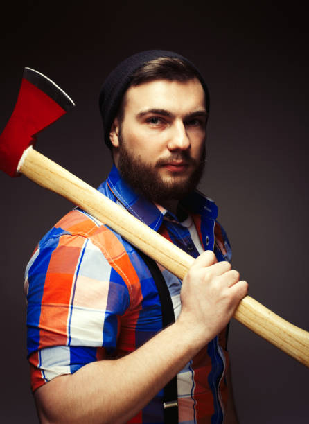 istock Young bearded man with big axe on shoulder standing against dark background 1384907926