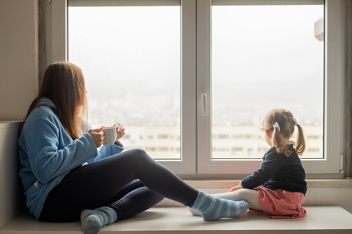 Mother and daughter looking out of the bedroom window on a rainy day