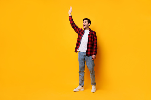 Hello, Nice to Meet You. Handsome excited young guy waving hand to the side free copy space and smiling on yellow studio background, greeting as notices someone walking, full body length banner