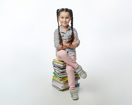 charming little girl with braided pigtails is sitting on a large stack of books.