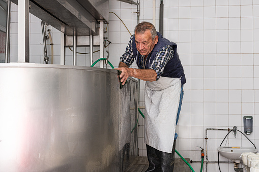 old man in an apron scrubbing the outside of stainless steel container. Factory