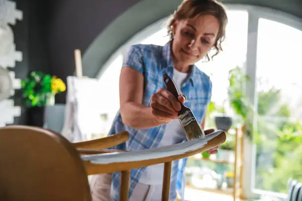 Low angle view of active mature woman repairing an old chair by painting it with white color during lockdown days at home.