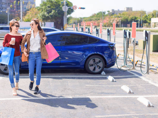 Happy  young woman standing on city parking near electric car, charging automobile battery from small city station, holding shopping bags and walking together stock photo