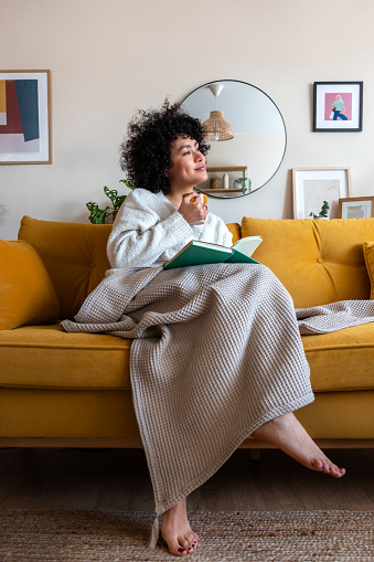 Pensive happy African american woman reading a book at home sitting on sofa relaxing drinking coffee. Vertical image. Lifestyle concept.