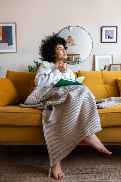 mulher afro-americana feliz pensativa lendo um livro em casa sentado no sofá relaxando bebendo café. imagem vertical - resting relaxation book break - fotografias e filmes do acervo