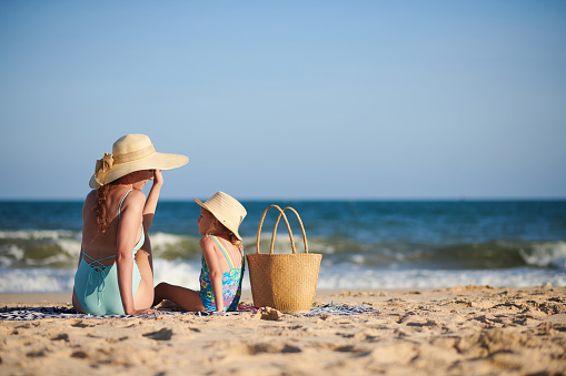 Mother and little daughter in straw hats sitting on beach, chatting and sunbathing