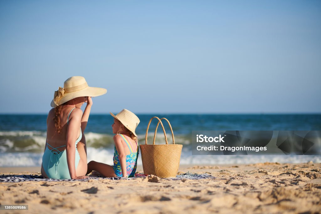 Mother and Daughter Sunbathing on Beach Mother and little daughter in straw hats sitting on beach, chatting and sunbathing Beach Stock Photo
