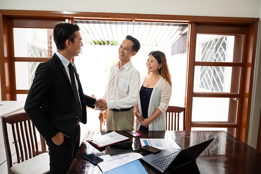 Happy male insurance agent shaking hand with an Asian couple after a successful real estate investment agreement signing.