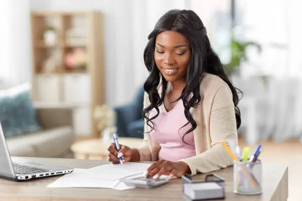 remote job, technology and people concept - happy smiling young african american woman with calculator and papers working at home office