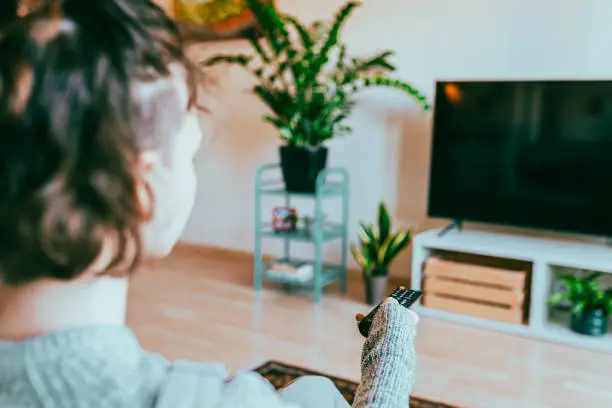 Photo of Back view of teenage girl sitting on sofa at home turning on TV with remote control