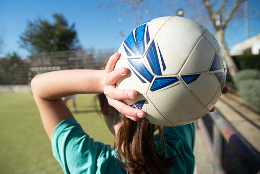 Close-up of teenage girls hands with ball. Back view of young girl with white and blue ball over her head on school stadium, ready to throw it to her friend. Womens football, healthy lifestyle concept
