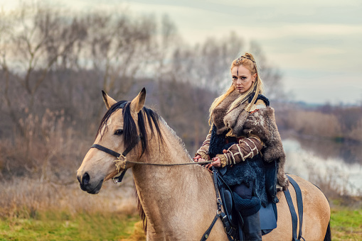 Beautiful Viking warrior royal female with braids and painted face riding horse in forest