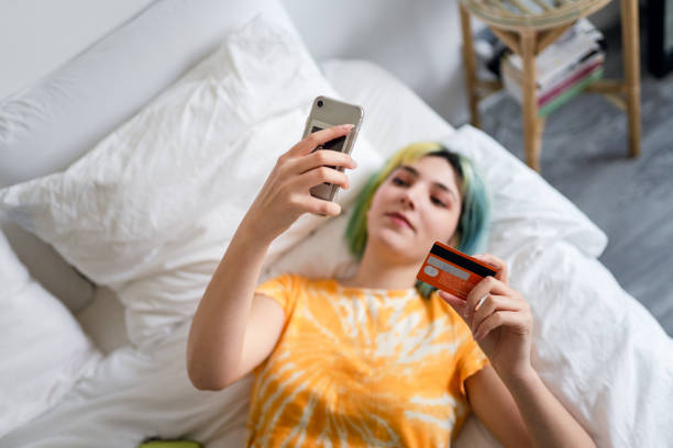 junge frau mit colorierten haaren kauft online mit einer kreditkarte ein - working bed smart phone bedroom stock-fotos und bilder