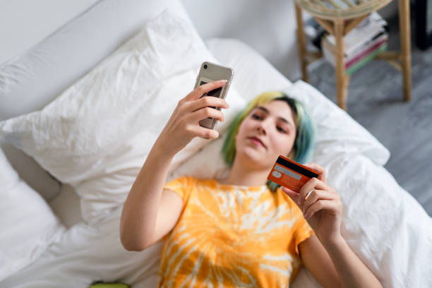 mujer joven con cabello teñido está comprando en línea con una tarjeta de crédito - working bed smart phone bedroom fotografías e imágenes de stock