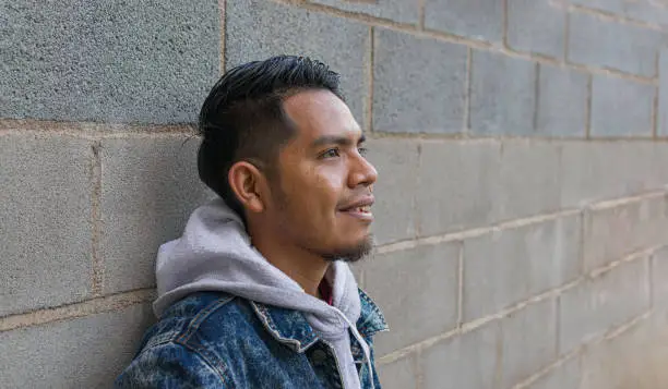 Photo of nice-haircut mexican man leaning on a wall smiling and looking away something