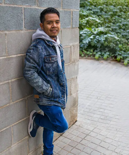 Photo of a street style simple guy with nice haircut leaning on a concrete bricks wall while looking at the camera