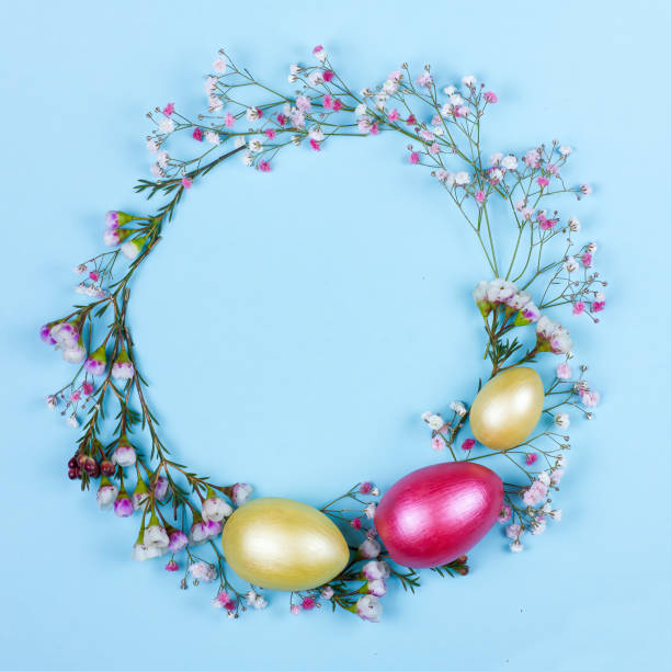 istock Easter wreath with spring flowers, green leaves and colored eggs on blue background. 1385071980