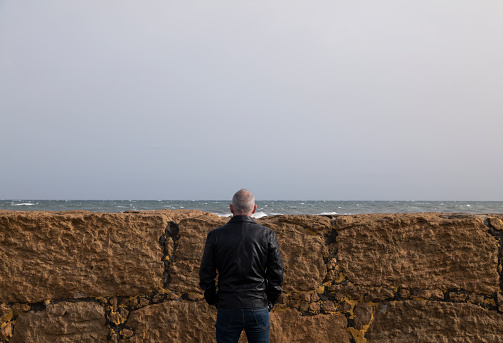 Rear view of adult man against short stone wall looking at sea