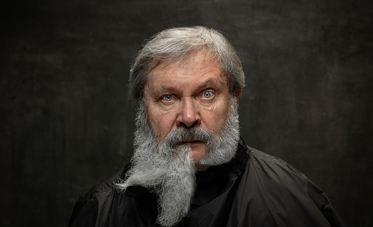 Vintage portrait of senior man with creative beard looking at camera isolated on dark vintage background. Concept of emotions, fashion, beauty, self-reinvention. Old man after visiting barbershop.