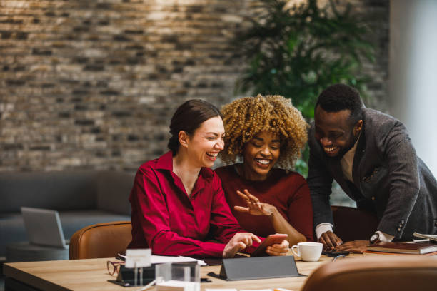 Three diverse business people hanging out and looking at funny memes Copy space shot of three diverse business colleagues hanging out in the office, having fun and laughing together at funny memes they are looking at on smart phone. teamwork red stock pictures, royalty-free photos & images