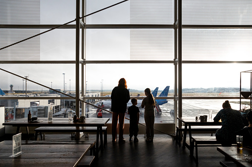 A rear-view shot of an unrecognizable mother and son and the boy's cousin standing in the airport terminal waiting to depart, they are looking at a plane out the airport window.