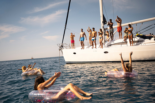 Young friends relaxing on floats and standing on sailboat in sea.
