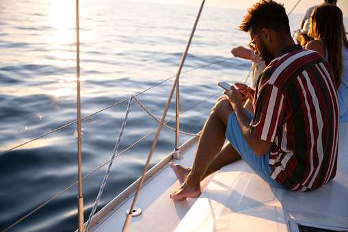 Young man sitting and using smart phone on sailboat in sea.