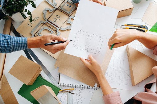 Top view of box designers desk. Man and woman hands at work. There is pencil pointing and blueprint scetch holding.